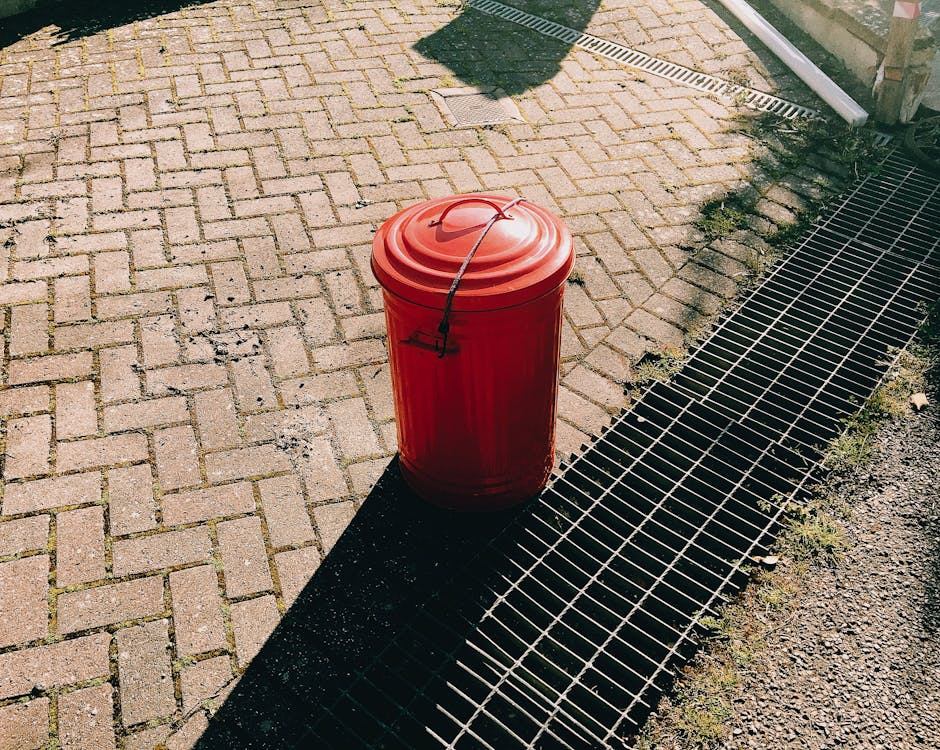 An image of a Red trash can on the pavement near sewer grates