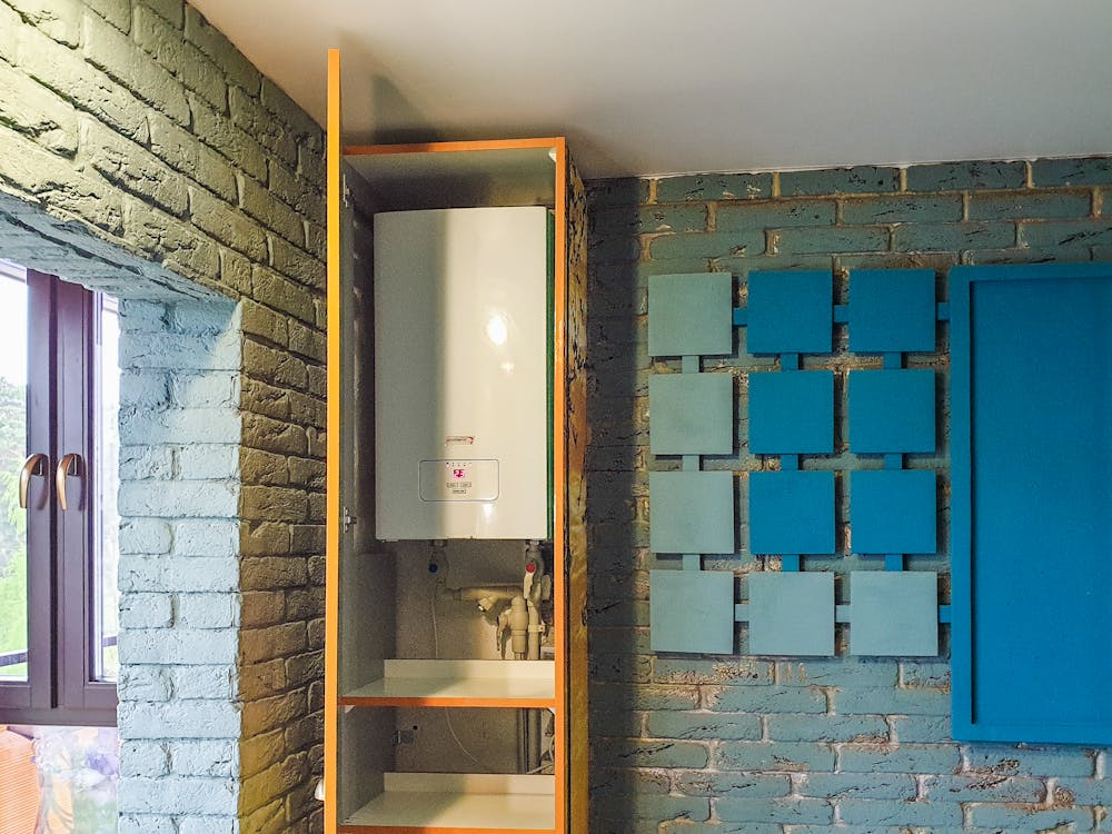 A modern white water heater is installed inside a wooden cabinet against a painted brick wall.