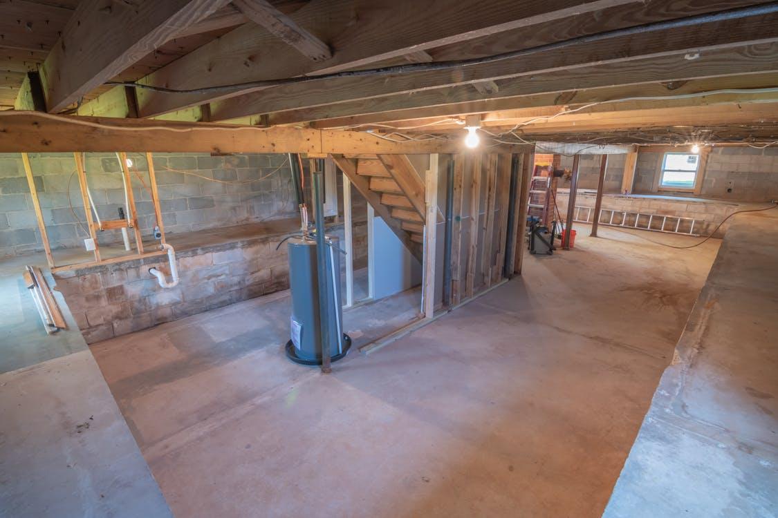 An unfinished basement with exposed wooden beams, a water heater installed near a staircase, and visible plumbing and electrical work.