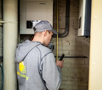 A technician in a gray hoodie and cap is inspecting or working on electrical equipment mounted on a brick wall.