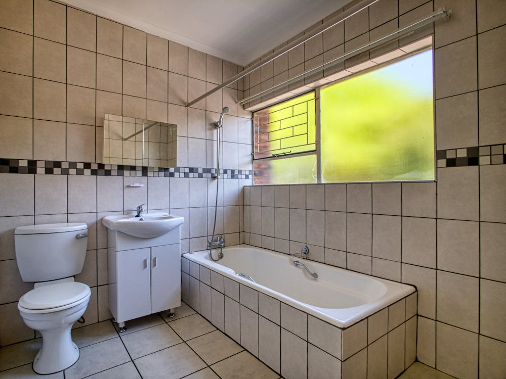 A tiled bathroom featuring a toilet, sink with a cabinet, bathtub with a handheld showerhead, and a large frosted window.