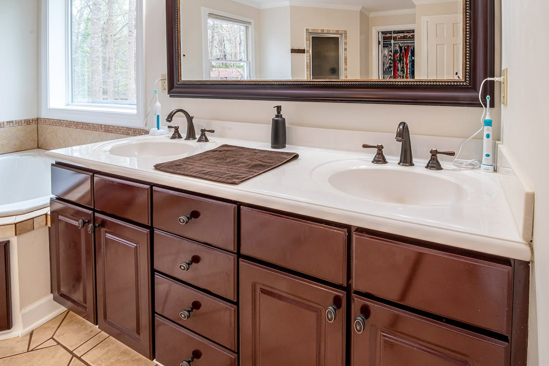 A bathroom vanity with a double sink, dark wood cabinets, a large framed mirror, and an electric toothbrush by the window.