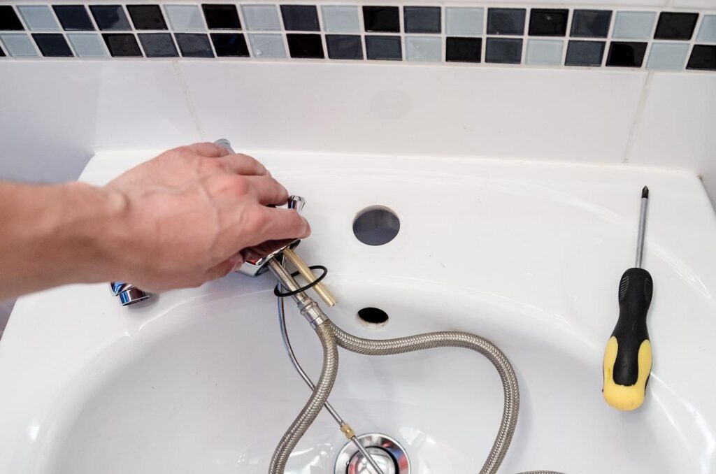 A close-up of a person installing a faucet on a bathroom sink with exposed supply lines and a screwdriver nearby.