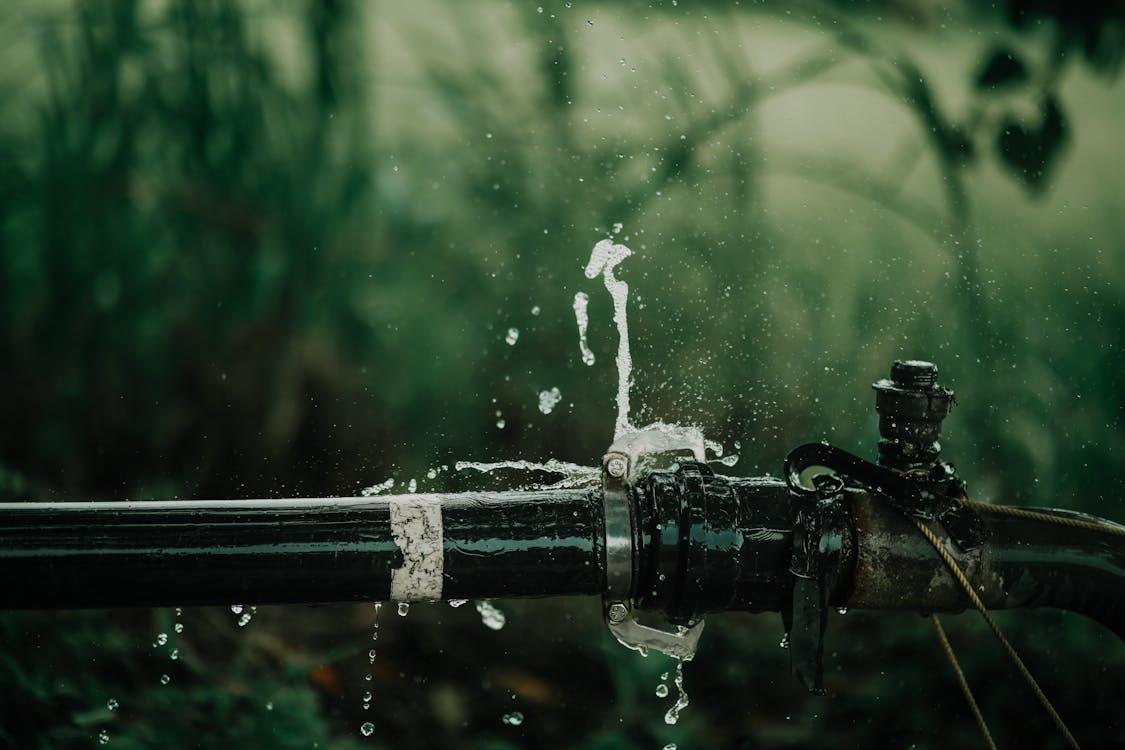 A close-up of a leaking black pipe outdoors, with water spraying out from a joint or crack, set against a blurred green natural background.