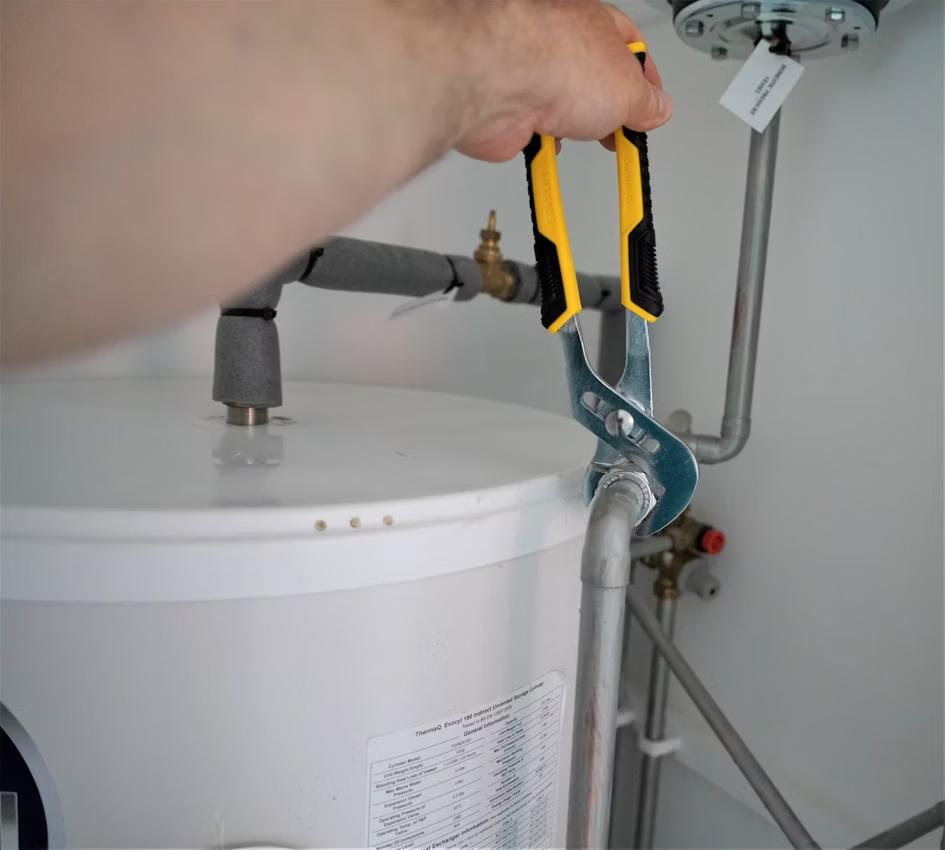 A person using adjustable pliers to tighten or loosen a pipe connection on a water heater.