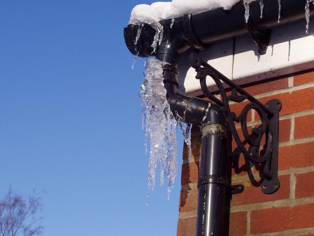 Frozen gutter and downspout on a brick house with icicles hanging, indicating cold winter weather and potential drainage blockage.