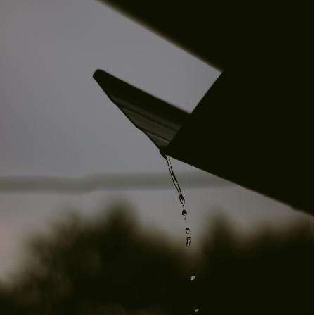 Close-up of rainwater dripping from the edge of a roof gutter during a dark, overcast day.