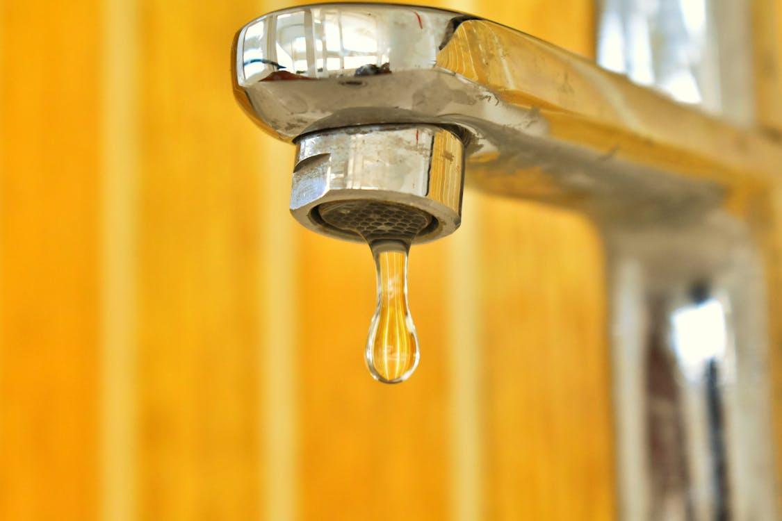  A close-up of a chrome faucet with a single droplet of water hanging from the spout against a yellow background.