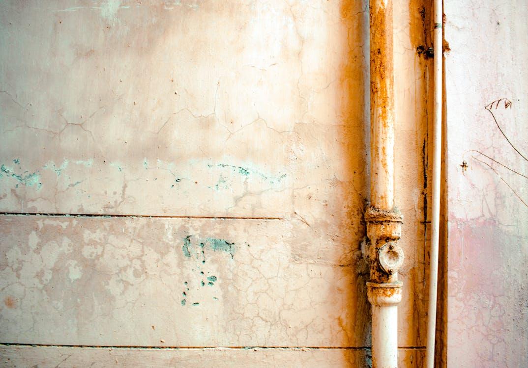 A rusted white pipe runs vertically along a cracked, weathered, and peeling pastel-colored wall.