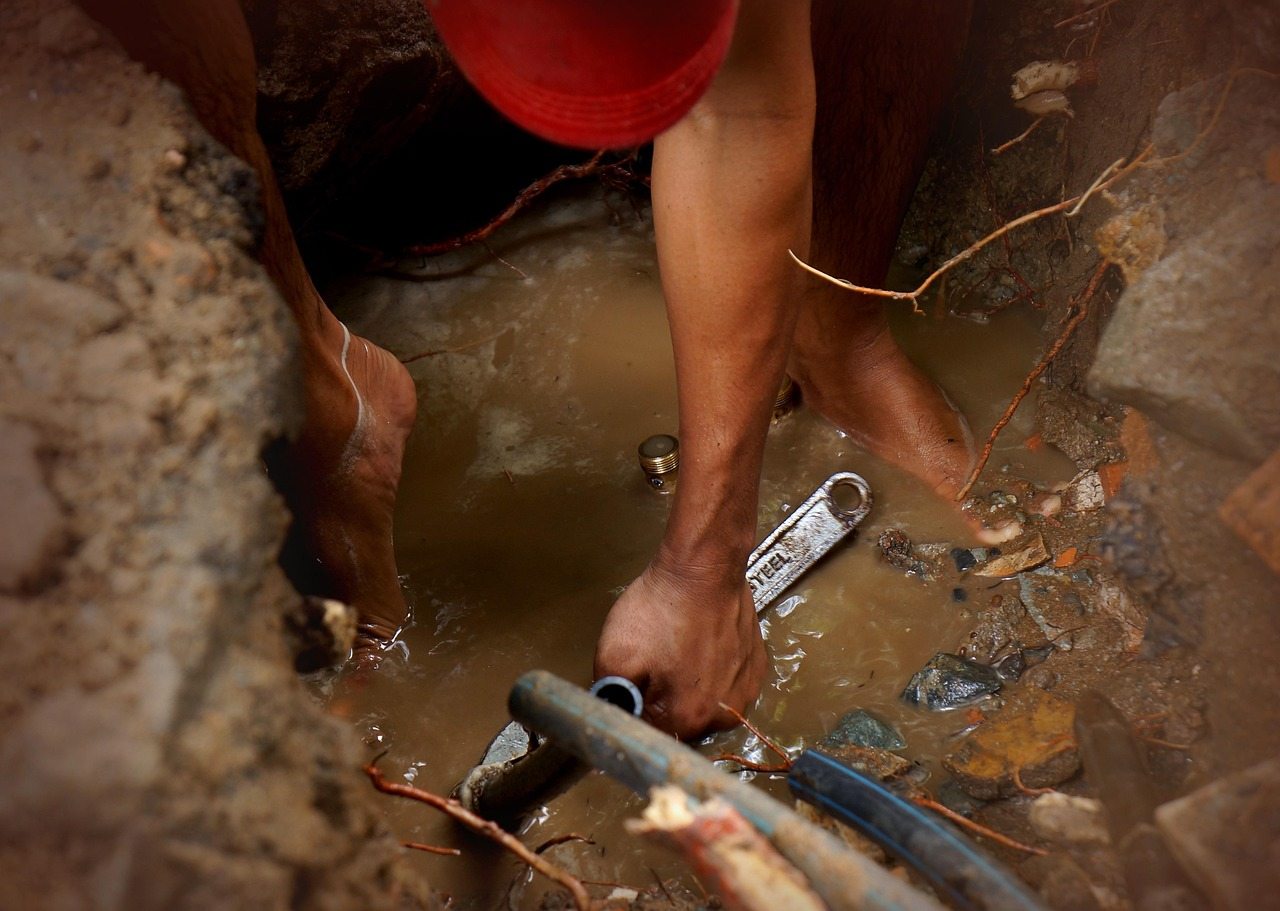 The image shows a person working barefoot in a muddy trench, possibly fixing plumbing, using a wrench.