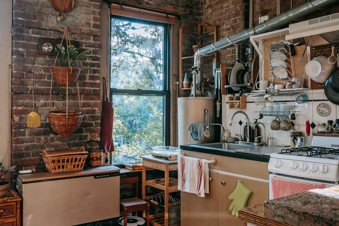 The image depicts a cozy kitchen with exposed brick walls, hanging plants, and a window that lets in natural light.