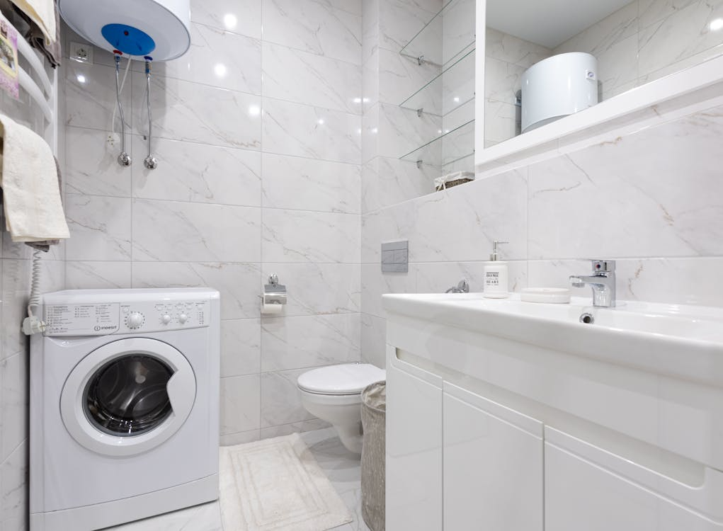 This image shows a modern, compact bathroom with a washing machine, toilet, and sink, featuring light marble tiles and minimalist design.