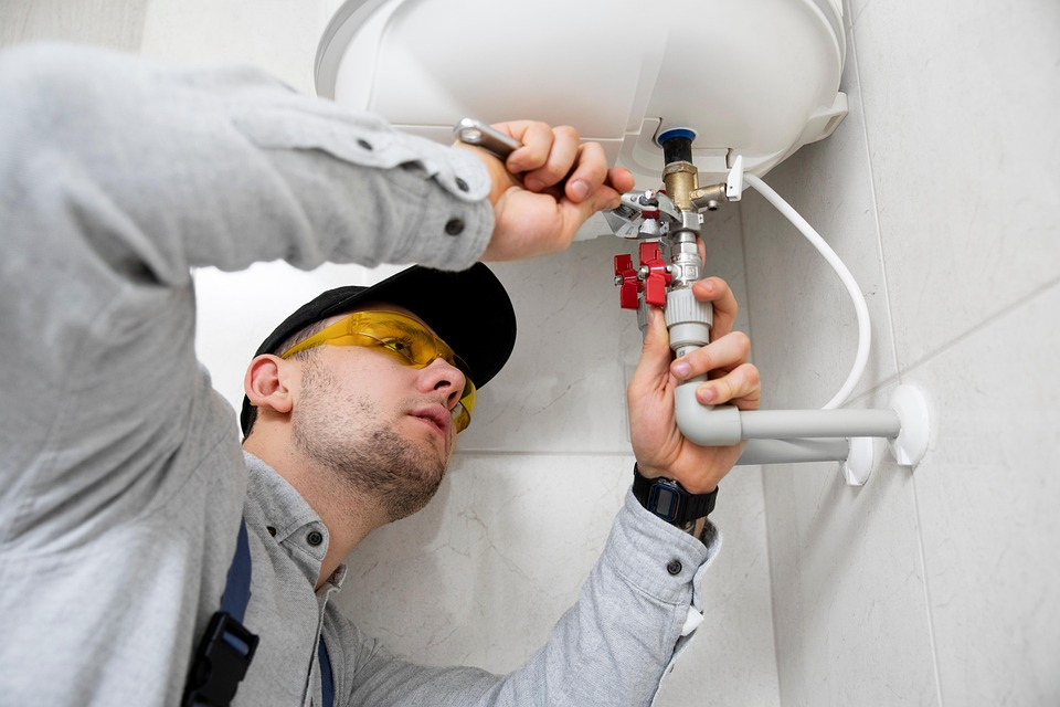 The image shows a plumber wearing safety glasses and using a wrench to work on a water heater or plumbing fixture mounted on a wall.
