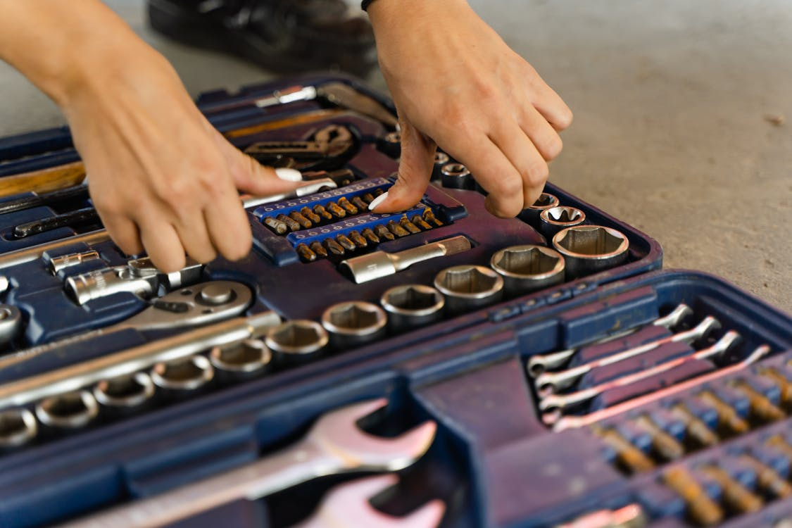 The image shows a close-up of hands organizing or selecting tools from a toolbox filled with various socket wrenches and bits.