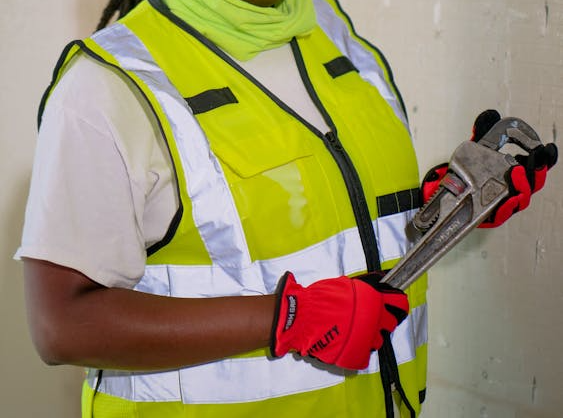A construction worker wearing a yellow hard hat, safety vest, and red gloves holds a large adjustable wrench while standing against a wall.