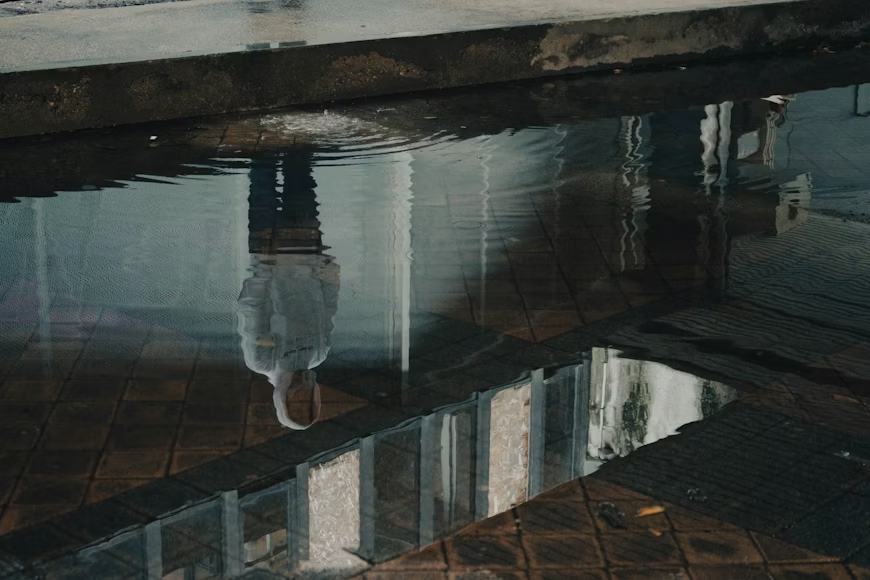 A reflective puddle shows a distorted upside-down image of a person standing near a tiled, wet surface.