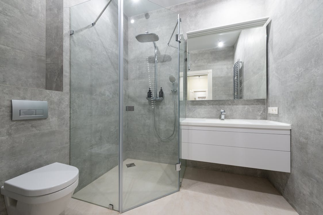 The image shows a sleek, modern bathroom with a walk-in shower, minimalist white vanity, and neutral gray tiles.