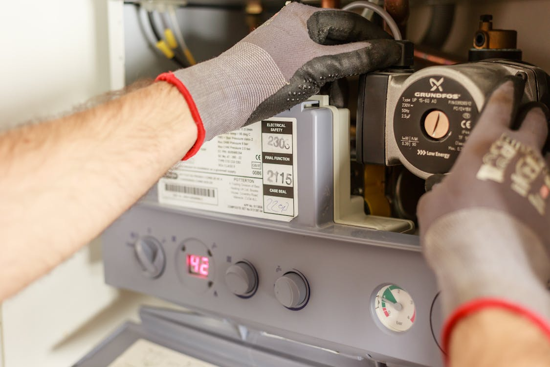 The image shows a close-up of a person's hand wearing a glove while working on a heating system or pump.