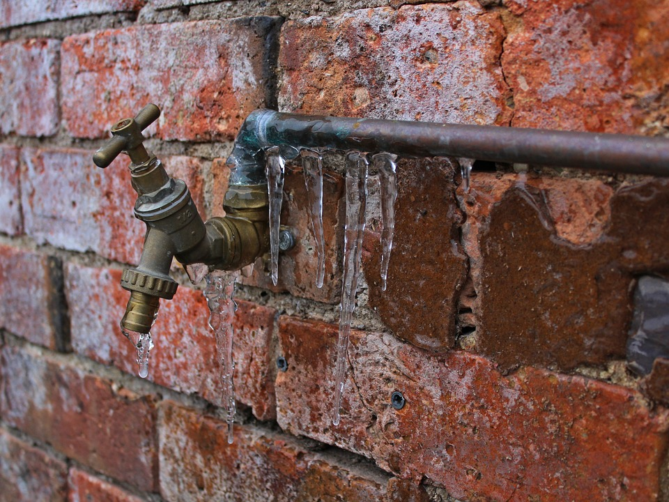 The image shows a water faucet on a brick wall with icicles forming around it, indicating freezing temperatures.