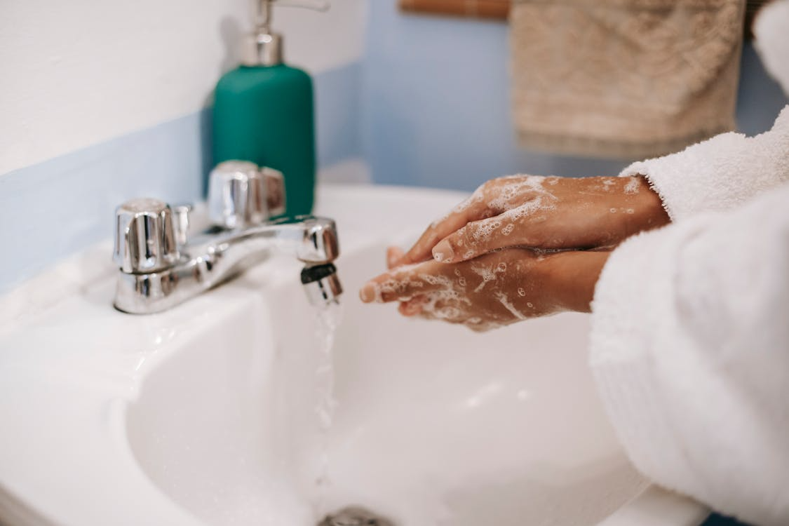 The image shows a person washing their hands with soap under a running faucet in a bathroom setting.