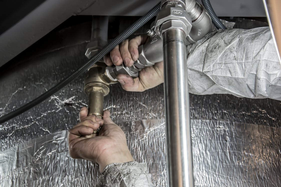The image shows a close-up of hands working on metal pipes, likely installing or repairing plumbing or HVAC systems.
