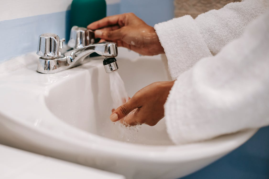 The image shows a person washing their hands under a faucet in a bathroom, wearing a white robe.