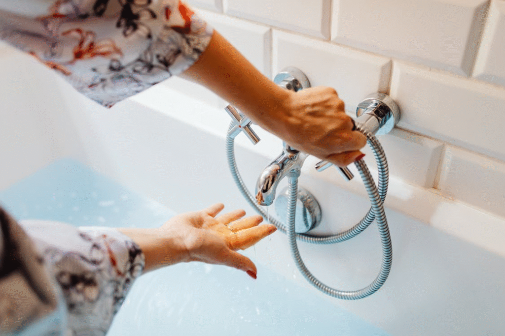 The image shows a person adjusting a handheld showerhead while water flows from it in a bathtub.