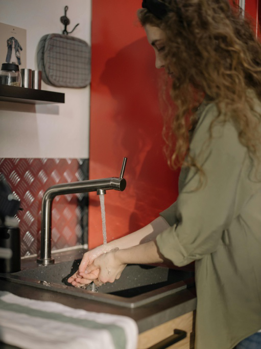 Woman in a brown long-sleeve shirt standing in front of a kitchen sink, checking water flow.