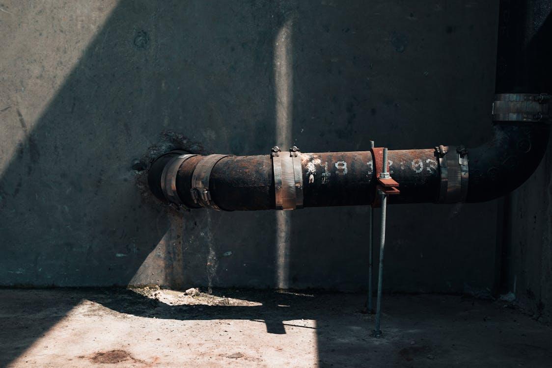 A rusted metal pipe secured with clamps runs horizontally along a concrete wall, partly lit by harsh sunlight.
