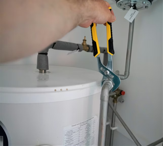 A hand uses adjustable pliers to tighten a metal pipe connection on a white water heater.