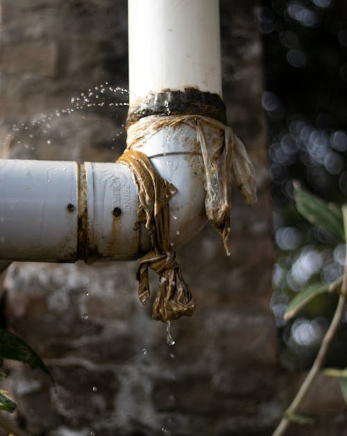 A leaky white PVC pipe joint wrapped with cloth or tape, spraying and dripping water outdoors near a brick wall.