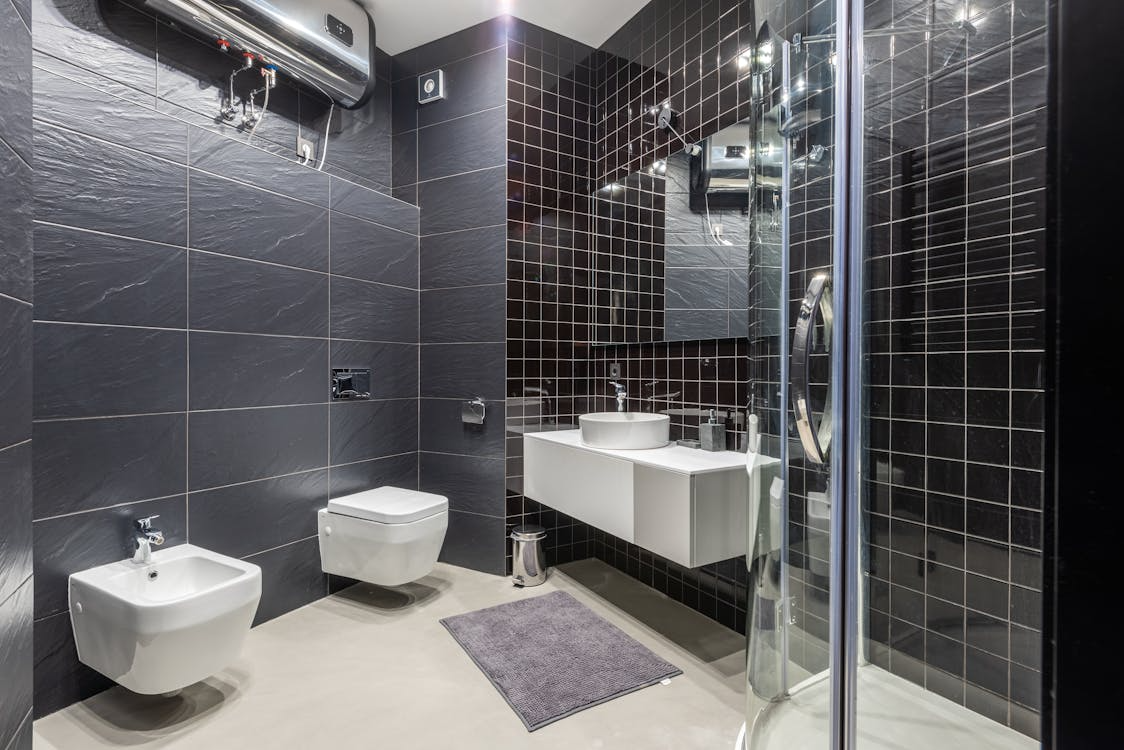 A modern, sleek bathroom with dark tiled walls, a glass shower enclosure, and minimalist white fixtures.