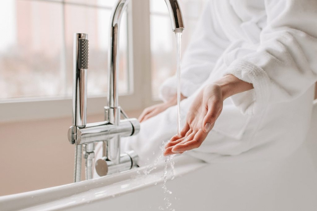 A person in a white bathrobe holds their hand under running water from a bathtub faucet.