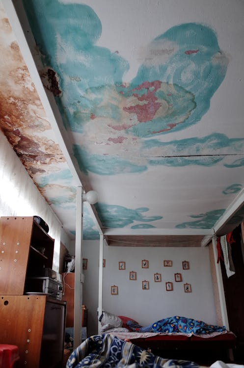 A small bedroom with a bed beneath a ceiling heavily stained and peeling from water damage and mold.