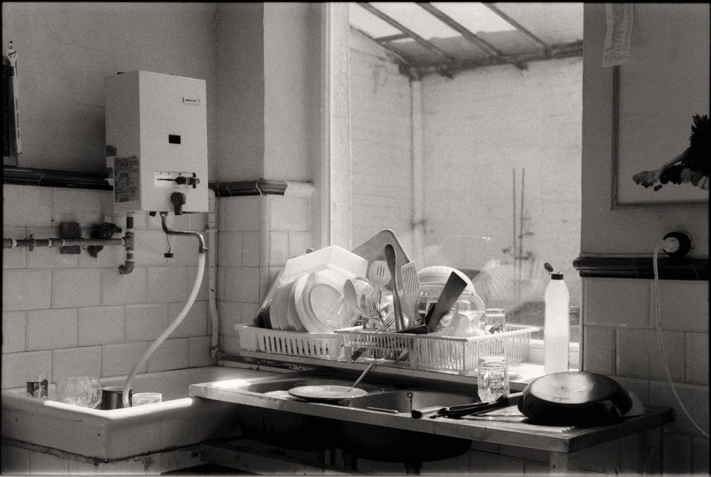 A black-and-white photo of a modest kitchen sink piled with drying dishes, lit softly by light from a window.